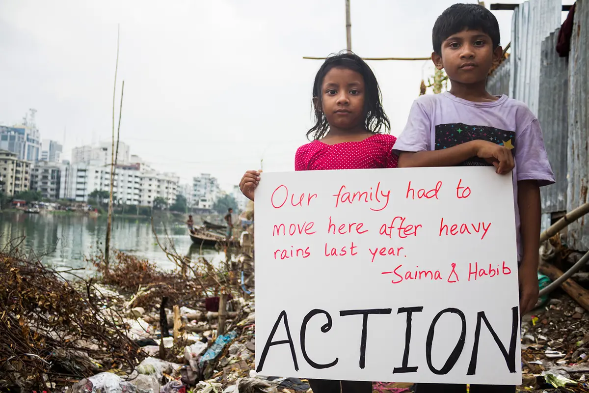 Two children pose in the slums with a sign reading "Our family had to move here after heavy rains last year - ACTION".